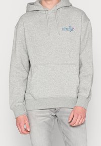 Sudadera gris con capucha, bolsillo frontal y logo con texto azul claro en el pecho izquierdo. Hecha de tela suave y texturizada con puños acanalados.