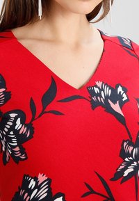 Femme portant un haut rouge à col en V avec de grands motifs floraux noirs, blancs et roses, et de longues boucles d'oreilles argentées à pampilles.