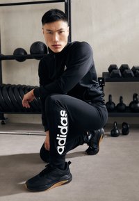 Black athletic outfit with zip jacket and fitted pants, featuring large white "adidas" logo. Black sneakers with gum soles. Gym background.