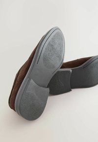 Chaussures en suède marron avec des semelles en caoutchouc gris, présentant un motif texturé et un design courbé au niveau du talon et de l'avant-pied, mettant en valeur une couture détaillée.