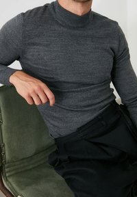 Grauer, langärmliger Rollkragenpullover aus strukturiertem Strickstoff, kombiniert mit hochtaillierten schwarzen Hosen, sitzend auf einem grünen Stuhl.