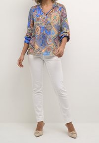 Blusa colorata con un motivo paisley in blu, beige e rosso, dotata di scollatura a V e maniche arrotolate, abbinata a jeans skinny bianchi.