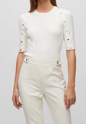 Femme portant un haut en maille côtelée blanc avec des boutons argentés sur les manches et un pantalon taille haute assorti avec des passants de ceinture à boutons.