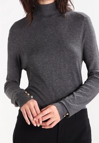 Pull à col roulé gris foncé en tissu doux avec une coupe ajustée. Présente des accents de boutons dorés sur les poignets pour plus de détail.