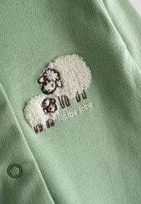 Hele roheline kangas, mis on tikitud valgete ja pruunide kasukatega lammastega ning allosas tikitud tekst "väike "