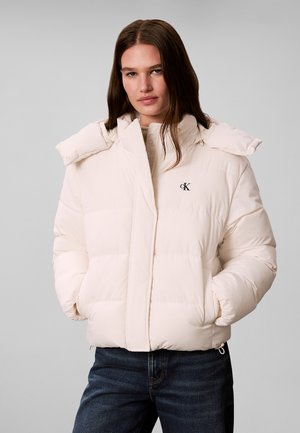 Femme portant une doudoune beige clair avec un col montant, les mains dans les poches, et un jean bleu foncé sur un fond gris uni.
