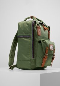 Zelený látkový batoh s hnědými koženými detaily, boční kapsou a vzorovaným zipem. Obsahuje polstrované popruhy a má pevný tvar.