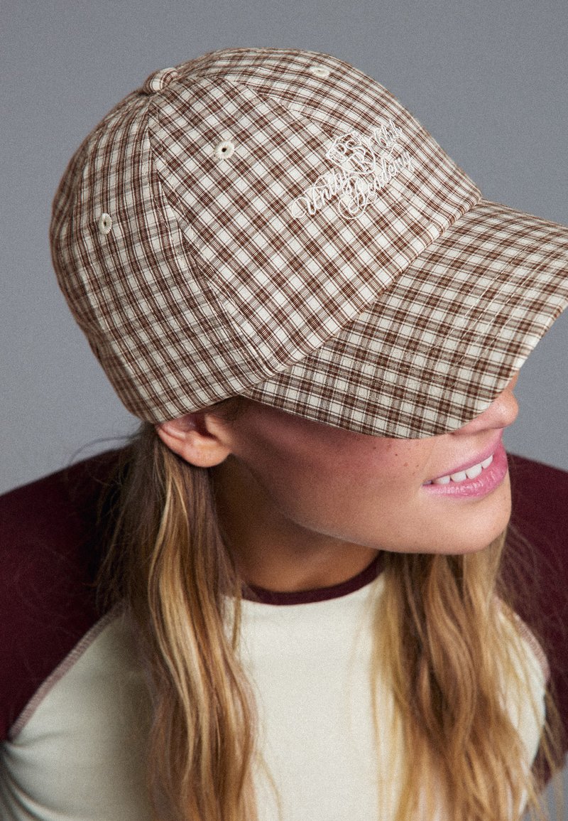 Femme portant une casquette à carreaux beige et marron avec un texte brodé, souriant et regardant légèrement sur le côté sur un fond uni.
