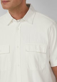Korte mouwen, geknopte blouse in het wit met subtiele verticale strepen. Beschikt over twee voorzakken en knoopsluitingen. Gladde stofstructuur.