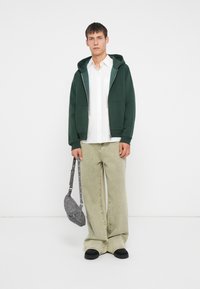 Zelena zip-up kapuca, bela majica, svetlozelene široke pantalone i crne cipele. Siva torba preko ramena sa zaobljenim oblikom i srebrnim detaljima.