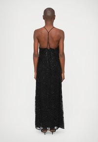 Pessoa de pé com as costas voltadas para o espectador, vestindo um vestido preto de lantejoulas sem mangas, com finas alças entrelaçadas.