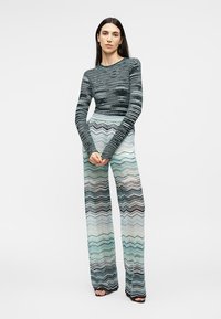 Žena stojaca so založenými rukami, oblečená v pruhovanom top s dlhým rukávom a širokých nohaviciach so vzorom cik-cak v odtieňoch zelenej, čiernej a bielej.