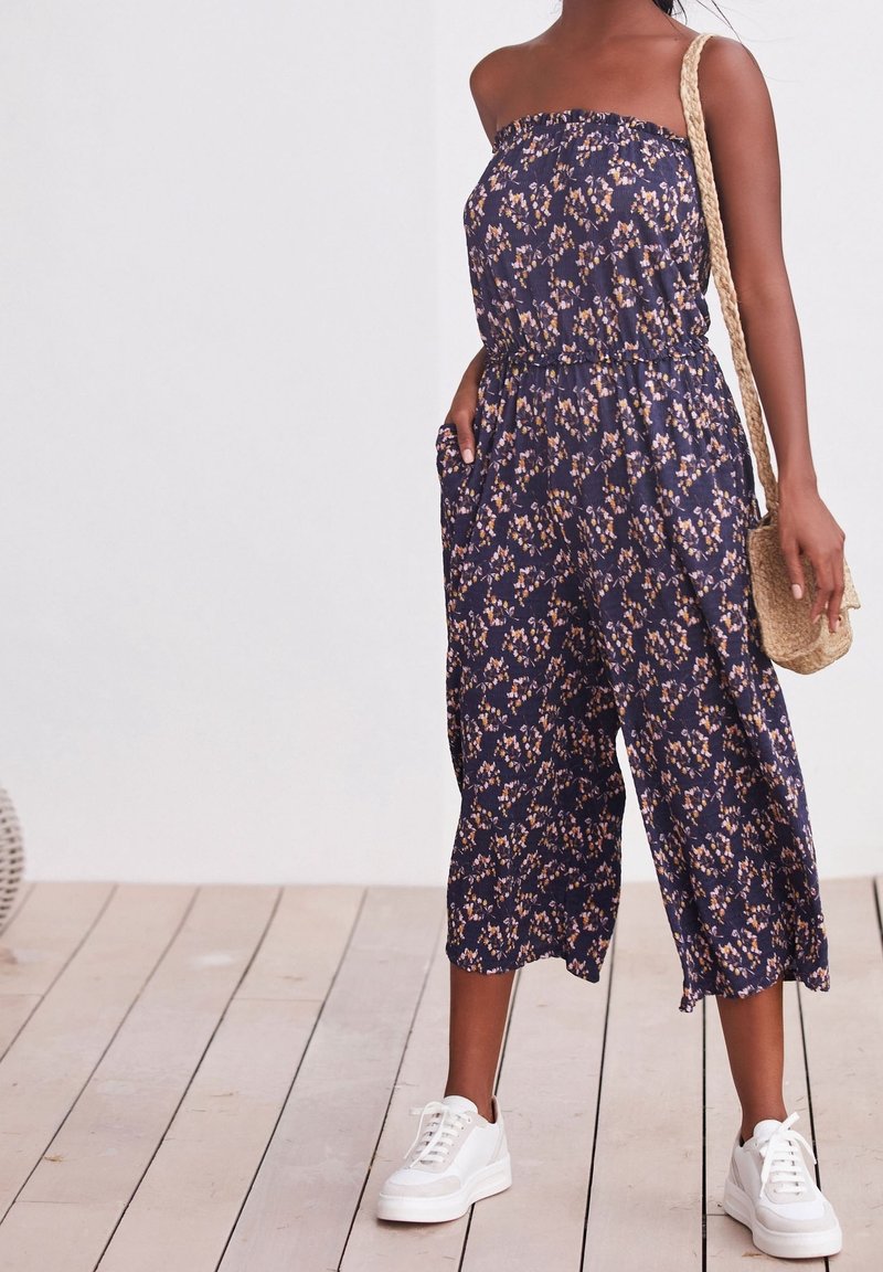 Námornícky květinový overal s elastickým vrškem, širokými nohavicemi a bočními kapsami, doplněný bílými teniskami a tkanou crossbody taškou.