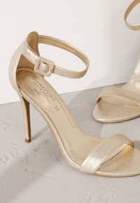 Sandale à talons hauts dorée avec bride à la cheville et bout ouvert, posée sur une surface texturée gris clair.