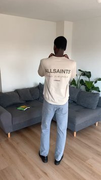 Άνδρας με την πλάτη, φοράει λευκό φούτερ AllSaints με κείμενο, ανοιχτόχρωμα τζιν και μαύρα παπούτσια σε ένα σαλόνι με γκρι καναπέ και πράσινο φυτό.