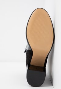 Semelle d'une chaussure noire à talon haut avec avant beige lisse et talon noir texturé, appuyée contre un mur blanc.