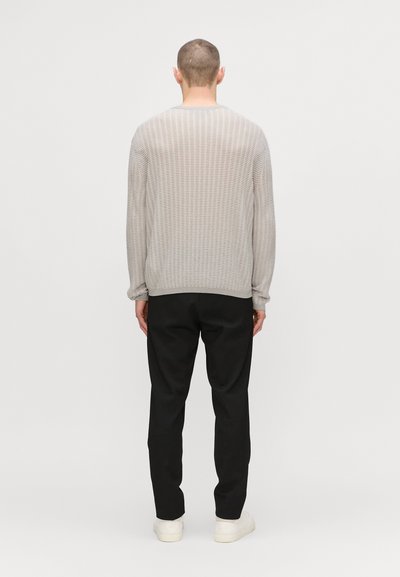 Pulover texturat de culoare gri deschis, cu mâneci lungi, design cu margini ondulate și gât rotund, asortat cu pantaloni negri și pantofi albi.