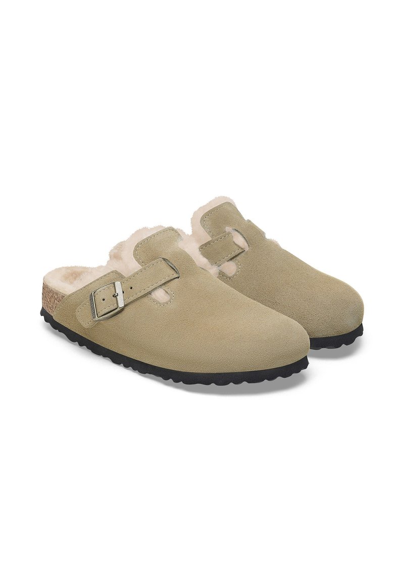 Sabots en daim beige avec détail de boucle, doublure intérieure douce et semelle en caoutchouc noir texturé. Bout rond et design sans arrière.