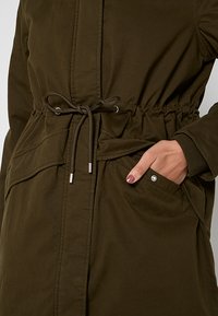 Olijfgroene katoenen jas met een trekkoord in de taille, grote zijzakken en zilveren hardwareaccenten voor sluiting en zakdetails.