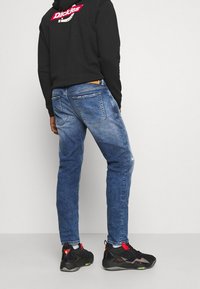 Blue denim jeans with a faded finish, black hoodie featuring a logo, and black sneakers with red accents. Visible stitching and back pockets.