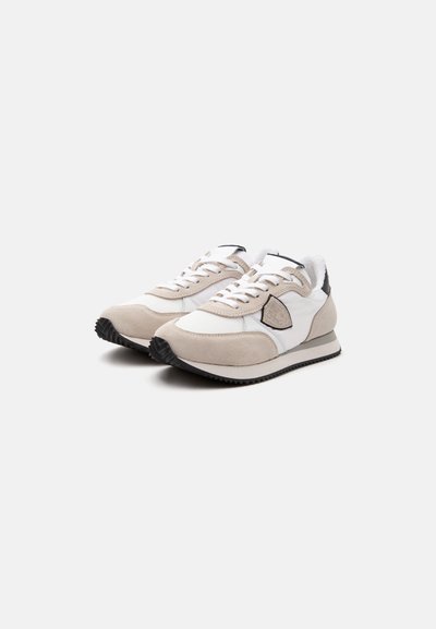 Paire de baskets basses blanches et beiges avec semelles noires et écusson avec logo sur les côtés, présentées sous un léger angle sur un fond uni.