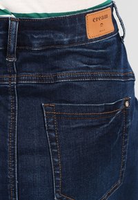 Tmavě modré džíny z denimu s oranžovým šitím, které mají koženou štítek s nápisem "cream" na pase a detail kapsy na zadní straně.