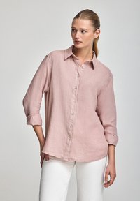 Camicia di lino rosa chiaro con maniche lunghe, chiusura a bottoni sul frontale e polsini arrotolati. Tessuto morbido, vestibilità sagomata e orlo curvo.