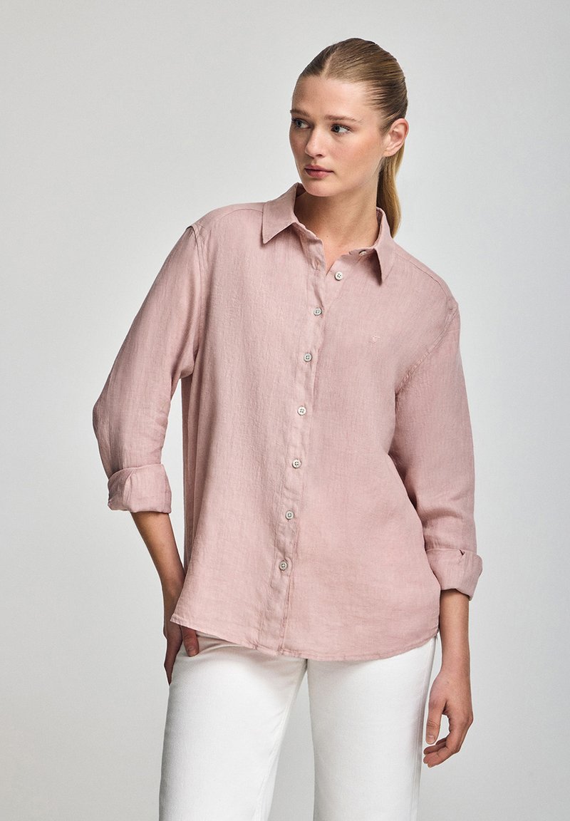 Camicia di lino rosa chiaro con maniche lunghe, chiusura a bottoni sul frontale e polsini arrotolati. Tessuto morbido, vestibilità sagomata e orlo curvo.