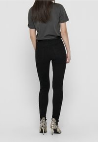 Schwarze, enganliegende Jeans mit hoher Taille und Rückentaschen, kombiniert mit einem grauen T-Shirt und gemusterten High-Heel-Stiefeln. Glatte Textur.