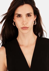 Žena s dlouhými tmavými vlasy a hnědýma očima, která má na sobě zlaté kruhové náušnice a černý top bez rukávů s hlubokým výstřihem do V.