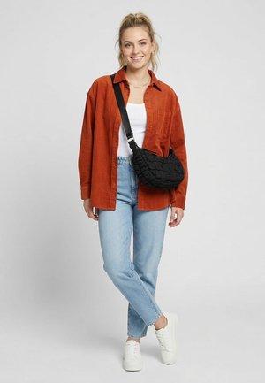 Junge Frau lächelnd, trägt ein rostfarbenes Hemd, weißes Tanktop, hellblaue Jeans, weiße Sneakers und trägt eine schwarze Schultertasche.