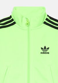 Fényes neon zöld Adidas dzseki fekete háromcsíkos részletekkel a vállakon, elülső cipzárral és hímzett fekete Adidas logóval a mellkason.