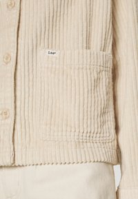 Beige corduroy jas met verticale ribbels, een voorklepzak en knopen. Het label "Lee" is zichtbaar vlakbij de zak.