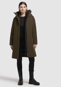 Donna che indossa un lungo cappotto in verde oliva con cappuccio, leggings neri e stivali neri alla caviglia, in piedi contro uno sfondo chiaro e semplice.