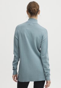 Svetlo modra pulover z visokim ovratnikom in dolgimi rokavi, s poudarjenima robovima in manšetami. Material se zdi mehak in teksturiran.