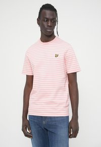 Ροζ κοντομάνικη μπλούζα με λευκές ρίγες, στρογγυλή λαιμόκοψη και μικρό κίτρινο λογότυπο πεταλούδας στο στήθος. Φορεμένη με μπλε τζιν.