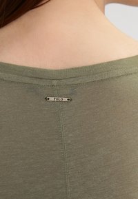 Olivově zelená košile s jemnou texturou, kulatým výstřihem a malou kovovou etiketou s nápisem "POLO" umístěnou na zadní části uprostřed.