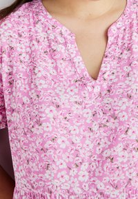 Růžové šaty s krátkými rukávy s květinovým vzorem zahrnujícím bílé a světle růžové květy, výstřihem do V a detaily s nařaseným lemem.