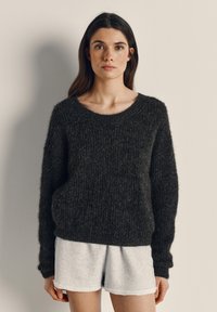 Frau mit langen dunklen Haaren, die einen strukturierten dunklen Pullover und hellgraue Shorts trägt, steht vor einer schlichten beigen Wand.