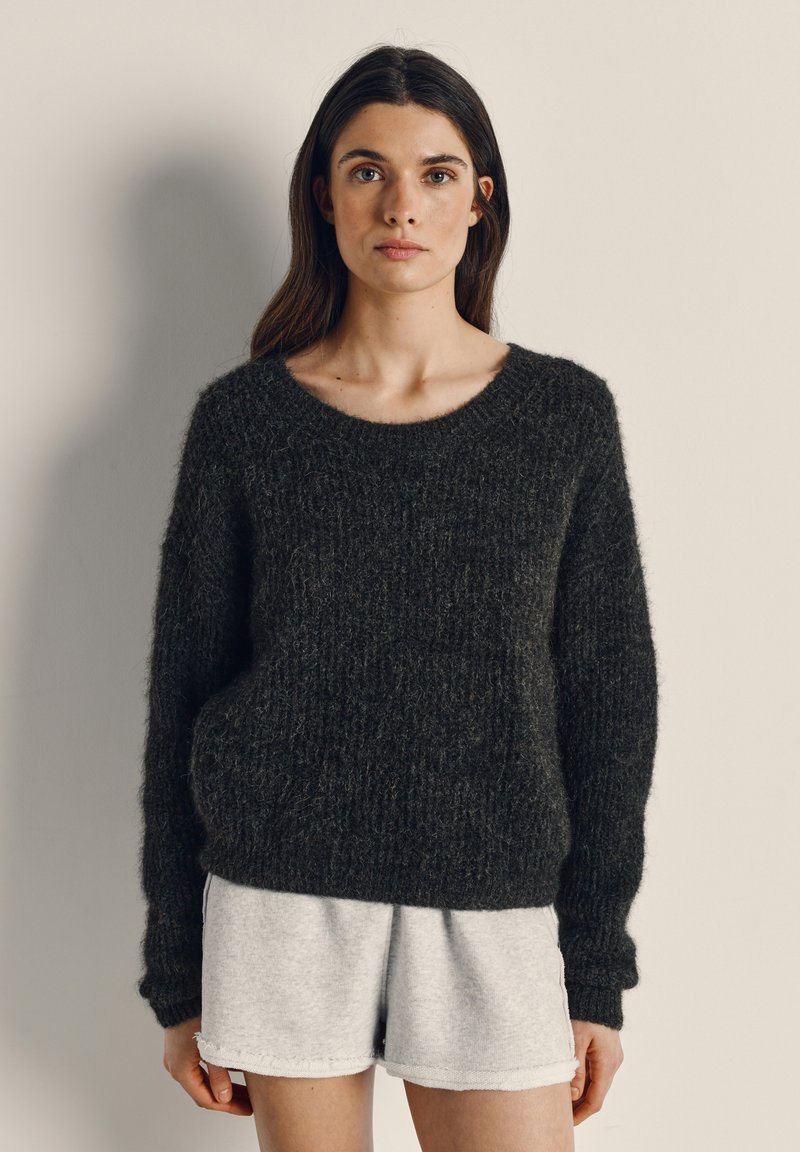 Frau mit langen dunklen Haaren, die einen strukturierten dunklen Pullover und hellgraue Shorts trägt, steht vor einer schlichten beigen Wand.