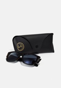 Ochelari de soare Ray-Ban cu ramă groasă din acoperire de țestoasă și lentile întunecate, așezați lângă un etui din piele texturată neagră cu un logo auriu.