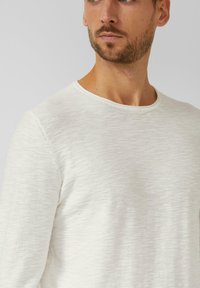 Man met een korte baard die een effen wit shirt met lange mouwen en ronde hals draagt, licht naar de zijkant gericht tegen een grijze achtergrond.