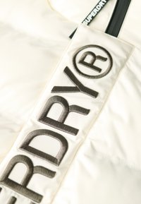 Cremefarvet polstret jakke med fremtrædende brune broderede logoer, en glat tekstur og en sort lynlås med en logo-tag.