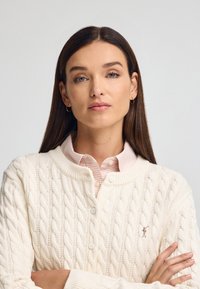 Weißer Strickcardigan mit rundem Kragen, versehen mit einem kleinen Logo. Über einem hellrosa Karohemd mit Kragen getragen. Glatte Textur, Knopfverschluss.