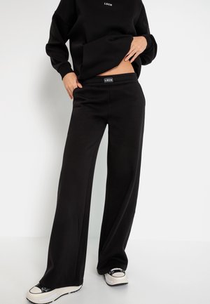 Personne portant un pantalon de survêtement noir à jambes larges et un sweat assorti, soulevant légèrement le t-shirt pour montrer la taille, associé à des baskets noires et blanches.