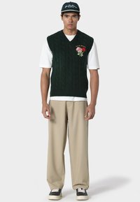 Vihreä neulottu hihaton liivi, jossa on palmikkokuvio ja kukka-ompelut; käytetty valkoisen t-paidan päällä, yhdistetty beigenvärisiin housuihin ja mustiin kenkiin.