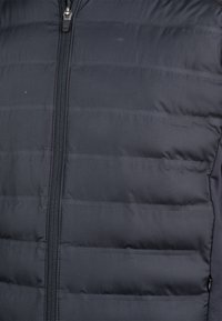 Gepolsterte schwarze Jacke mit vertikalen Steppnähten und einem Reißverschluss vorne. Glattes Material mit glänzender Oberfläche und subtilen Nähdetails.