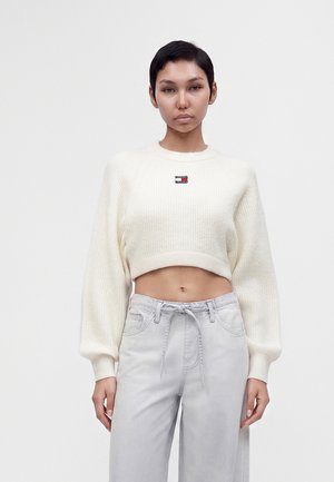 Femme aux cheveux courts et foncés portant un pull court blanc et un jean gris clair taille haute sur un fond uni.