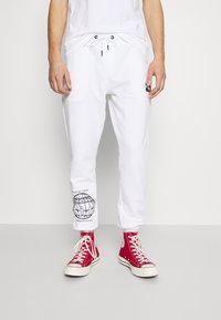 Vita joggarebyxor med elastiskt midjeband och dragsko, med en svart grafisk tryck på vänster ben. Tillsammans med röda högaxlade sneakers.