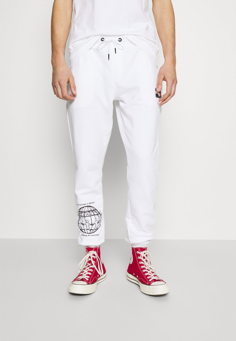 Vita joggarebyxor med elastiskt midjeband och dragsko, med en svart grafisk tryck på vänster ben. Tillsammans med röda högaxlade sneakers.